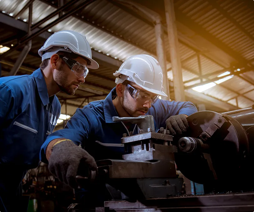 Two engineers in hard hats inspecting machined hardware on a shop floor.